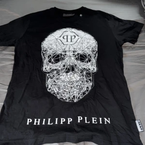 Svart t-shirt med döskalle från Philipp Plein - Cool svart t-shirt från Philipp Plein med stor vit grafisk döskalle på framsidan och logotyptryck. T-shirten har rund hals och är tillverkad i mjuk bomull. Perfekt för dig som gillar statement-plagg och streetwear. Betalning genom Swish 