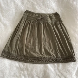 Olivgrön kjol från h&m - Supersöt olivgrön kjol från h&m med knytband i midjan och broderad kant längst ner. Kjolen har en lätt utställd form och är gjord i mjuk bomull, perfekt för en avslappnad stil. Midja: 33cm, Längd: 50cm