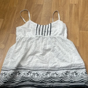 Vit och svart prickig linnetopp från H&M - Säljer en söt vit linnetopp från H&M med svarta prickar och blommigt mönster längst ner. Toppen har tunna axelband och dekorativa veck framtill. Perfekt för varma dagar och enkel att matcha med jeans eller kjol.