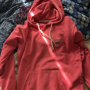 Rosa hoodie från Gant - Säljer en snygg rosa hoodie från Gant med broderad logga på bröstet. Tröjan har huva med dragsko och en stor magficka framtill. Perfekt för en avslappnad stil.