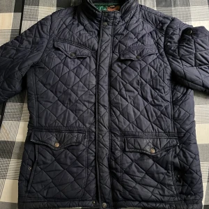 Svart quiltad jacka från Reset - Säljer en svart quiltad jacka från Reset med hög krage och fyra fickor framtill med knappstängning. Jackan har dragkedja och tryckknappar samt ett grönt innerfoder. Perfekt för kyliga dagar.