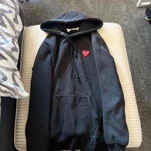 Svart hoodie med rött hjärta från Comme des Garçons Play - Svart hoodie med huva och dragsko från Comme des Garçons Play. På bröstet finns ett broderat rött hjärta med ögon. Tröjan har magficka och ribbade muddar vid ärmslut och nederkant. Perfekt för en avslappnad stil. Skriv vid frågor😎