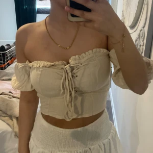 Beige offshoulder croppad topp med snörning - Säljer en beige offshoulder topp med croppad passform och snörning framtill. Toppen har volangdetaljer vid ärmar och halsringning, samt en smockad rygg för extra komfort. Perfekt för sommardagar!