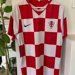 Kroatiens landslagströja från Nike! L  - Säljer en klassisk Kroatien fotbollströja från Nike med rödvita schackrutor, blå Nike-logga och landslagsemblem på bröstet. Tröjan är kortärmad och har röda detaljer på ärmarna samt blå inslag på sidorna. Perfekt för fotbollsfans! Tröjan är i finfint skick! 