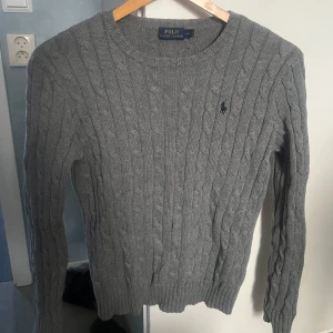 Grå stickad tröja från Polo Ralph Lauren - Säljer en klassisk grå stickad tröja från Polo Ralph Lauren med kabelstickat mönster och rund halsringning. Tröjan har lång ärm och den ikoniska broderade loggan på bröstet.