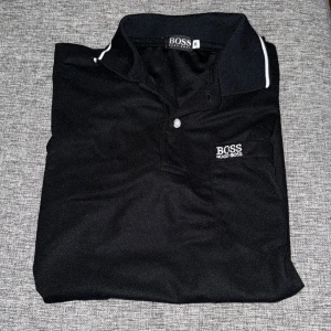 Svart pikétröja från Hugo Boss - Klassisk svart pikétröja från Hugo Boss med vit logga broderad på bröstet och vit detalj på kragen. Tröjan har korta ärmar och knäppning framtill. Perfekt för en stilren och sportig look.