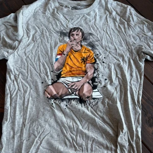 Smoking Cruyff T-shirt - Snygg grå t-shirt från Nakatá Football Club med coolt tryck av en tecknad fotbollsspelare i orange tröja på framsidan. Klassisk passform och rund hals. Tillverkad i mjuk bomull, perfekt för dig som gillar sportig stil.
