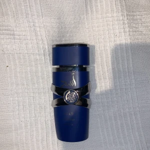 Asad Eau de Parfum från Lattafa - Snygg parfymflaska i matt mörkblått med silverdetaljer och arabisk text. Flaskan har en unik form med en dekorativ silverring och ett blått emblem i mitten. Sprayfunktion och matchande blått lock. Perfekt för dig som gillar exklusiv design och orientaliska dofter.