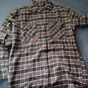 Brun och svart rutig skjorta från bikbok - Snygg rutig skjorta i brunt och svart med vita linjer. Skjortan har två bröstfickor med lock, klassisk krage och knäppning framtill. Perfekt för lager-på-lager och chill stil. Långärmad och i mjukt material som känns skönt mot huden.