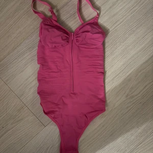 Rosa bodysuit med dragkedja - Säljer en rosa bodysuit med smala axelband och snygg dragkedja framtill. Toppen har en v-ringad urringning och lätt rynkade kupor för en feminin look. Perfekt att styla med jeans eller kjol för en trendig outfit.