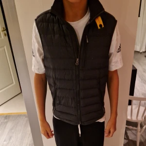 Svart dunväst från Parajumpers - Säljer en svart dunväst från Parajumpers i ungdomsstorlek M. Västen har quiltad design, hög krage och dragkedja framtill. Två praktiska fickor med dragkedja och en snygg gul detalj vid halsen. Perfekt för lager-på-lager under kyliga dagar.