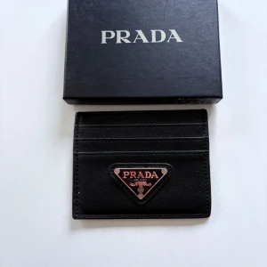Svart Prada plånbok med logga - Säljer en stilren svart plånbok från Prada med ikonisk triangelformad metallogo framtill. Plånboken har flera kortfack och är tillverkad i tåligt syntetmaterial. Kommer med originalbox, dustbag och äkthetskort. Perfekt accessoar för dig som gillar lyxiga detaljer.