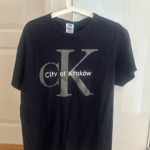 Mörkblå t-shirt City of Kraków JHK - Snygg mörkblå t-shirt från JHK i 100% bomull med stort tryck 'City of Kraków' och en stor silverfärgad bokstav K på bröstet. Klassisk rund hals och korta ärmar, perfekt för dig som gillar enkel men stilren design.