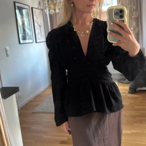 Svart omlottblus med puffärm - Supersnygg svart blus med omlottdesign och djup v-ringning. Blusen har puffiga långärmar, smockad rygg och snygg rynkning framtill som ger en cool siluett. Perfekt att styla med smycken och kjol för en trendig look.