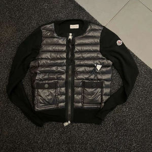 Svart dunväst från Moncler - Snygg svart dunväst från Moncler med quiltad framsida, två stora fickor med knappar och dragkedja framtill. Västen har Moncler-logga på ärmen och är tillverkad i en mix av dun och stickat material för en cool kontrast.