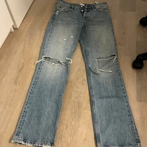 Blå ripped jeans från Zara - Säljer ett par blå jeans från Zara med raka ben och snygga slitningar vid knäna. Jeansen har hög midja, klassisk femficksdesign och knappar framtill. 