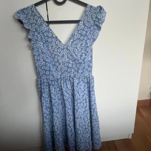 Blommig blå kortklänning från SHEIN - Supersöt blå klänning från SHEIN med vitt blommönster, volangdetaljer vid axlarna och v-ringad hals. Klänningen har omlottknytning i ryggen och är tillverkad i mjuk polyester. Perfekt för sommarens alla tillfällen, lätt och flowy passform.