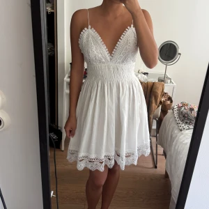 Vit spetsklänning från Zara - Supersöt vit kortklänning från Zara med tunna axelband, v-ringad spets upptill och smockad midja. Kjolen är lätt och har broderad spetskant. Öppen rygg och romantiska detaljer gör den perfekt för sommarens alla tillfällen. Materialet är bomull.