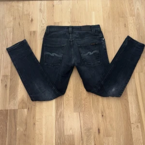 Svarta jeans från Nudie Jeans, 31/32 - Säljer ett par svarta jeans från Nudie Jeans i storlek 31/32. Jeansen har klassisk femficksdesign, snygga slitningar och Nudie-loggan på bakfickorna. Modellen är rak med normal passform och tillverkad i mjukt bomullstyg med lite stretch.
