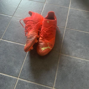 Röda fotbollsskor från Puma - Snygga röda fotbollsskor från Puma med gula detaljer och logga på ovansidan. Skorna har snörning, platt sula med dobbar och är tillverkade i syntetmaterial för bästa grepp på planen. Perfekta för dig som vill sticka ut på matchen!