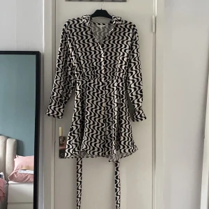 Svartvit mönstrad kortklänning - Kort klänning med svartvitt vågigt mönster, långärmad och med knappar framtill. Klänningen har krage och ett matchande bälte i midjan för snygg passform. Perfekt för dig som gillar statement-plagg med cool retrovibe.