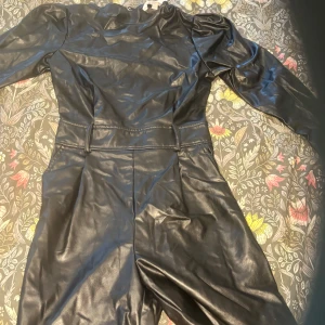 Svart skinnklänning från H&M - Cool svart klänning i skinnimitation från H&M med långa ärmar och markerad midja. Klänningen har en stilren look med lätt puff vid axlarna och rund hals. Perfekt för dig som vill sticka ut med en edgy vibe.