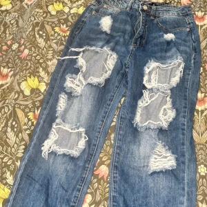Blå ripped jeans från Missguided - Säljer ett par blå jeans från Missguided Petite med stora slitningar och råa hål på båda benen. Jeansen har klassisk femficksdesign, dragkedja och knapp framtill. Modellen är rak och har en avslappnad passform. Perfekta för en edgy streetstyle-look.