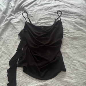 Svart linnetopp från Bik Bok - Säljer en svart linnetopp från Bik Bok med tunna axelband och draperad framsida. Toppen har en elegant, asymmetrisk volangdetalj på ena sidan och är tillverkad i ett mjukt, slätt material som faller snyggt. Perfekt för dig som gillar stilrena och trendiga plagg.