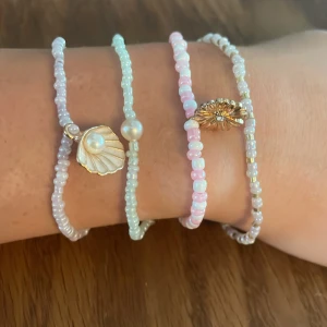 Pärlarmband kitt med 4 armband - Fyra handgjorda armband med små pärlor i pastellfärger. Ett armband har ljusblå pärlor och en vit pärla, ett har vita och guldiga pärlor, ett har rosa och vita pärlor med en guldig blomberlock, och ett har ljuslila pärlor med snäckberlock.
