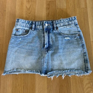 Jeanskjol med slitningar  - Säljer en kort jeanskjol från Zara i ljusblå tvätt med rå kant och slitna detaljer både fram och bak. Kjolen har klassiska fickor, bälteshällor och knappstängning. Perfekt för dig som gillar en edgy och avslappnad stil.