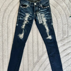 Affliction Raquel skinny jeans blå - Mörkblå skinny jeans från Affliction, modell Raquel. Snygga slitningar framtill och på bakfickorna, markerade kontrastsömmar och nitar vid fickorna. Jeansen har låg midja och är tillverkade i stretchig bomullsdenim för en tight passform.