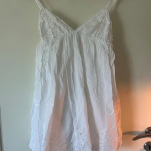 Vit kort klänning från Zara - Supersöt vit kort klänning/ lång top från Zara med tunna axelband och broderade detaljer längst ner. Klänningen har en v-ringad front och är tillverkad i 100% bomull, vilket gör den lätt och skön. Perfekt för varma dagar och sommarhäng med vänner.