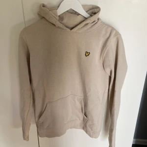 Beige hoodie från Lyle & Scott Junior - Snygg beige hoodie från Lyle & Scott Junior med klassisk gul logga på bröstet. Tröjan har huva, känguruficka och är tillverkad i mjuk bomull. Perfekt för en chill och stilren look.