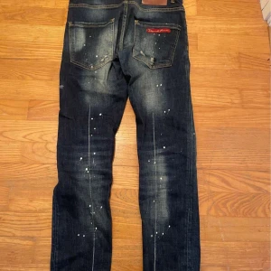 Dsquared2 mörkblå jeans med stänk - Säljer ett par snygga Dsquared2 jeans i mörkblå tvätt med coola vita färgstänk och slitningar.  Dsquared2-logga vid knappen och bakfickan. Jeansen har raka ben. 