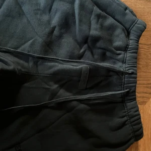 Svarta mjukisbyxor shorts Essentials FOG SS22 - Svarta mjukisbyxor från Fear of God Essentials med elastisk midja och dragsko. Byxorna har diskret logga framtill och är tillverkade i mjuk bomullsmix för maximal komfort. Perfekta för en chill och stilren streetwear-look.