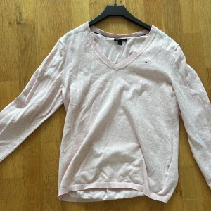 Ljusrosa v-ringad tröja Tommy Hilfiger - Ljusrosa långärmad tröja från Tommy Hilfiger med v-ringning och diskret logga på bröstet. Tröjan är i mjuk bomull och har en klassisk, normal passform. Perfekt för dig som gillar stilrena och enkla plagg med en touch av preppy.
