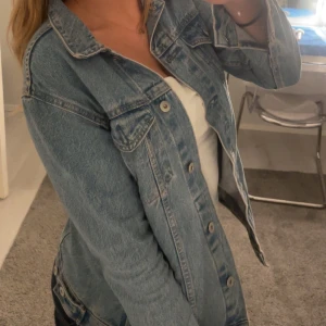 Klassisk ljusblå jeansjacka - Säljer en snygg ljusblå jeansjacka med klassisk passform och silverfärgade knappar. Jackan har två bröstfickor med lock och knäppning framtill. Perfekt att slänga över en hoodie eller t-shirt för en chill look.