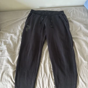 Svarta mjukisbyxor Peak Performance M - Svarta mjukisbyxor från Peak Performance med ribbade muddar vid benslut, snörning i midjan och två sidofickor. Snyggt logomärke på ena benet. Perfekta för chill eller träning, tillverkade i mjukt bomullsmaterial för extra komfort.