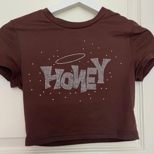Brun croppad topp med strass från SHEIN - Croppad brun topp från SHEIN med korta ärmar och coolt strassmönster framtill där det står 'HONEY' och en glittrig gloria ovanför. T-shirten är i mjuk bomull och har en figurnära passform som sitter snyggt till höga jeans eller kjol.
