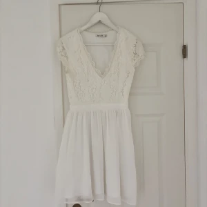 Vit spetsklänning från Nly One - Supersöt vit kortklänning från Nly One med spets upptill och v-ringad hals. Kjolen är lätt och flödande, medan överdelen har fina spetsdetaljer och korta ärmar. Perfekt för student eller sommarens alla tillfällen!
