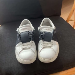 Valentino Garavani Open sneakers vit/blå - Snygga Valentino Garavani Open sneakers i vitt skinn med bred mörkblå läderdetalj över snörningen. Klassisk rund tå, platt sula och diskreta nitar baktill. Perfekta för dig som gillar exklusiva och stilrena sneakers med en twist. De ser lite sämre ut i verkligheten än på bilden för de är lite mer slitningar på tån och i hälen på insidan av skon som ej syns på bilden