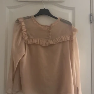 Beige blus med volangdetaljer - Supersöt beige blus i transparent material med långa ärmar. Blusen har volangdetaljer över axlar och bröst samt små knappar framtill. Perfekt för dig som gillar romantisk och feminin stil. Från h&m