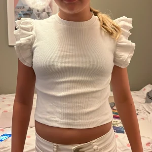 Vit ribbad topp med volangärmar Gina Tricot - Supersöt vit ribbad topp från Gina Tricot med korta volangärmar som ger en extra touch. Toppen är croppad och har rund halsringning. Perfekt för dig som gillar en stilren men ändå lekfull look.