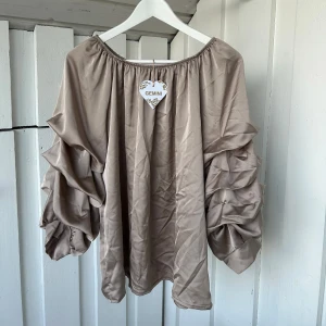 Beige blus med volangärmar från Gemini - Säljer en beige blus från Gemini med bred resår i halsen och riktigt snygga volangärmar som ger en cool och unik look. Materialet är mjukt och har ett lätt glansigt utseende, perfekt för dig som gillar statement-ärmar och vill sticka ut lite extra.