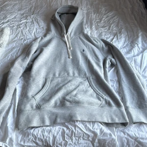 Grå hoodie från Polo Ralph Lauren - Säljer en grå hoodie från Polo Ralph Lauren med klassisk broderad logga i mörkblått på bröstet. Tröjan har huva med snörning, stor magficka och långa ärmar. Perfekt för chill dagar och enkel att matcha med jeans eller joggers.