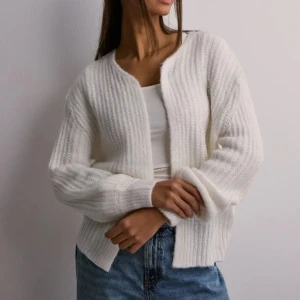 Vit stickad kofta - Säljer en vit stickad kofta med ribbad struktur och långärmade ärmar. Koftan har en loose passform och är öppen framtill utan knappar eller dragkedja. Perfekt att slänga över en t-shirt eller linne för en chill look. Materialet känns mjukt och fluffigt.