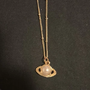 Guldigt halsband med planetberlock - Unikt halsband i guldton med en cool planetberlock där en vit pärla utgör planeten och en glittrig ring omger den. Kedjan har små detaljer längs länkarna som ger extra stilpoäng. Perfekt för dig som gillar rymdinspirerade smycken och vill sticka ut.
