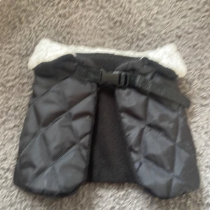 Svart quiltad täcke till sin käpphäst med fuskpäls - Svart quiltad dunväst till sina käpphästar med mjuk vit fuskpäls vid kragen och justerbart bälte med spänne framtill. Utsidan är i syntetmaterial och insidan har fleece för extra värme. Perfekt för kyliga dagar när du vill hålla stilen enkel men mysig.