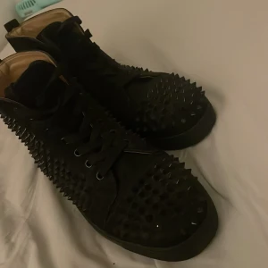 Svarta Louboutin sneakers med nitar - Grymma svarta sneakers från Louboutin med mattsvarta nitar över hela skon och den ikoniska röda sulan. Skorna har snörning och är tillverkade i mocka och skinn, vilket ger en lyxig känsla. Perfekta för dig som vill sticka ut med en edgy vibe. Dom e äkta fick dom av min storebror har tuvärr kvitto och box 