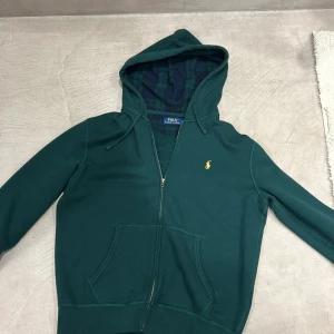 Grön hoodie från Polo Ralph Lauren - Snygg mörkgrön hoodie från Polo Ralph Lauren med gul broderad logga på bröstet. Tröjan har dragkedja, huva med snörning och praktiska fickor framtill. Insidan av huvan har ett rutigt mönster i grönt och blått. Perfekt för chill dagar.